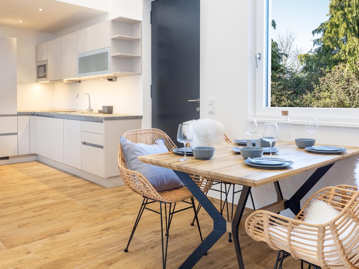 Helles Appartement mit Küchennische und Sitzgelegenheit. Das Fenster eröffnet den Blick auf einen Wald.