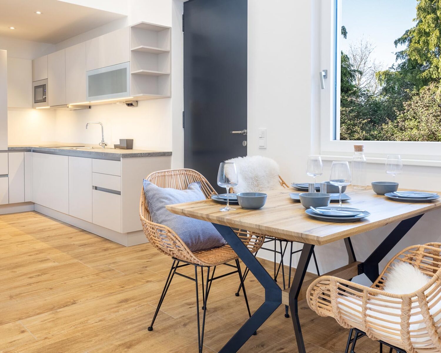 Helles Appartement mit Küchennische und Sitzgelegenheit. Das Fenster eröffnet den Blick auf einen Wald.